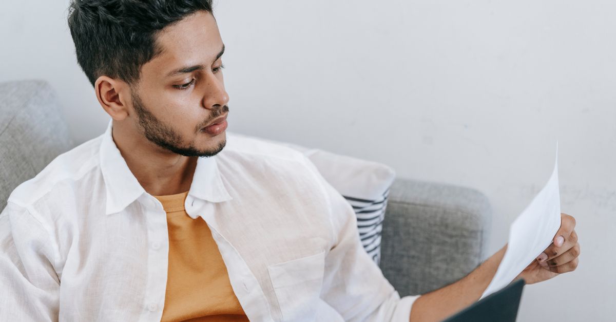 A young man sitting on a sofa reading a printed document closely