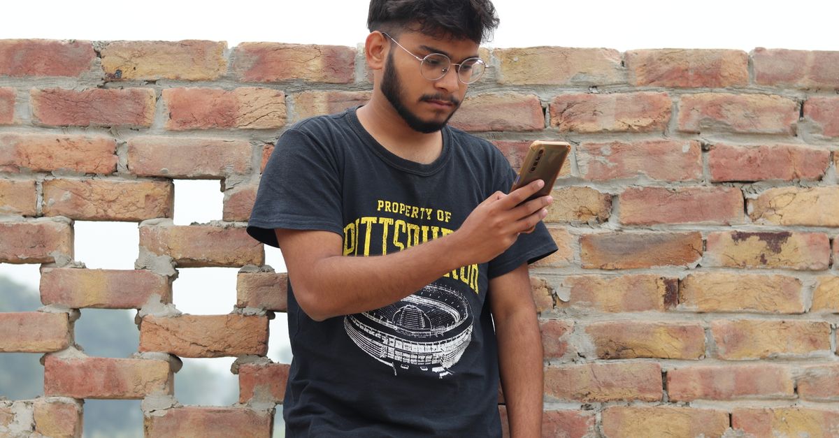 Indian man in glasses looking carefully at his phone in front of a brick wall