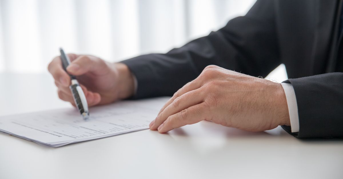 Hands in a formal suit holding a pen over a printed document on a desk