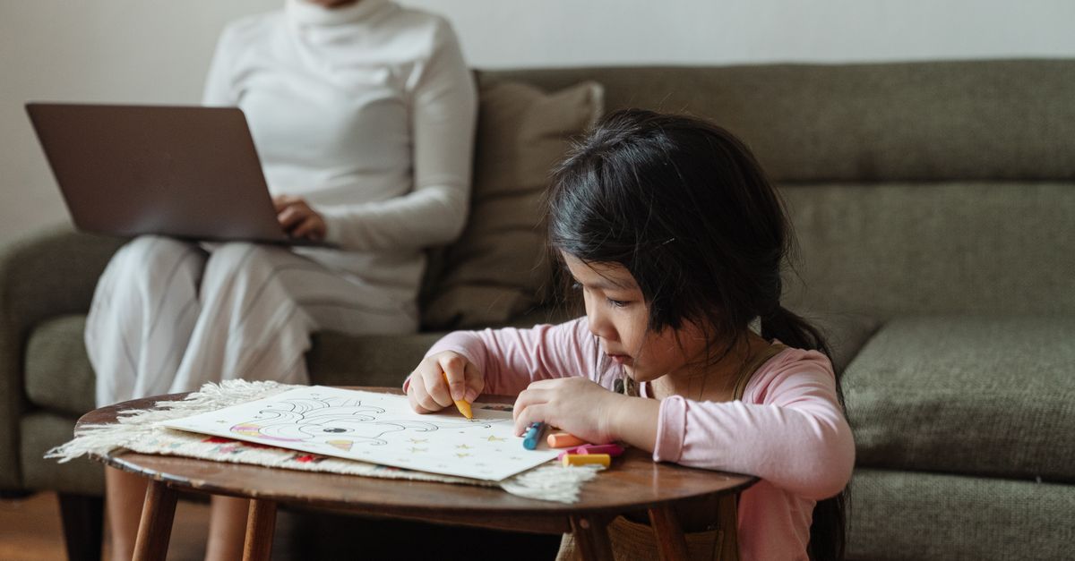 Homemaker working on a laptop from the sofa while her young child draws nearby.