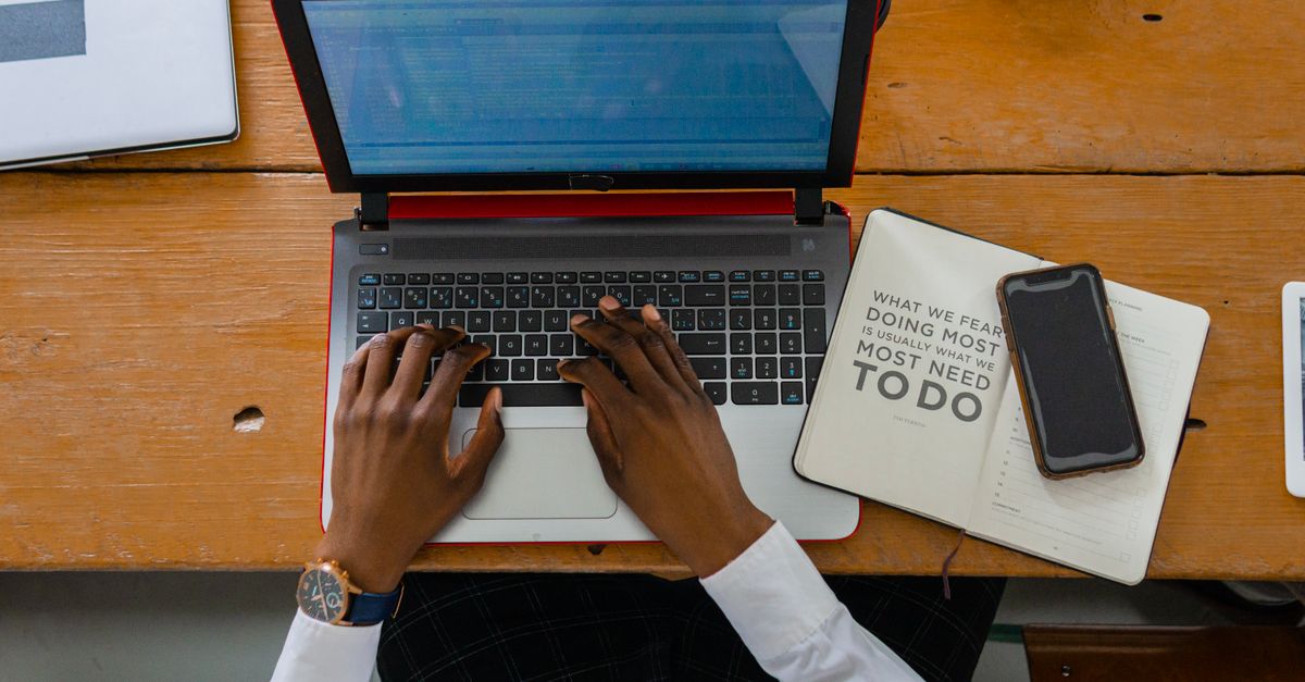 Hands typing on a laptop on a wooden desk with a notebook and a phone beside it